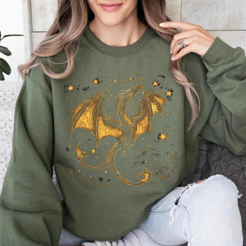 May include: Olive green sweatshirt featuring a golden dragon design with outstretched wings and star accents. The dragon is intricately detailed, with a textured appearance. The design is centered on the front of the sweatshirt, creating a fantasy-themed aesthetic.
