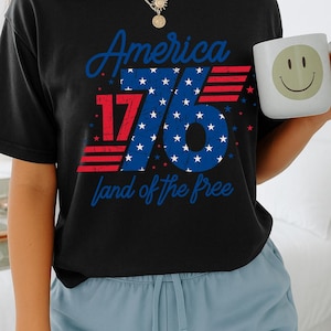 May include: Black t-shirt with the text "America 1776 land of the free" in red and blue. A white mug with a smiley face is held in the hand. The person is wearing blue shorts.