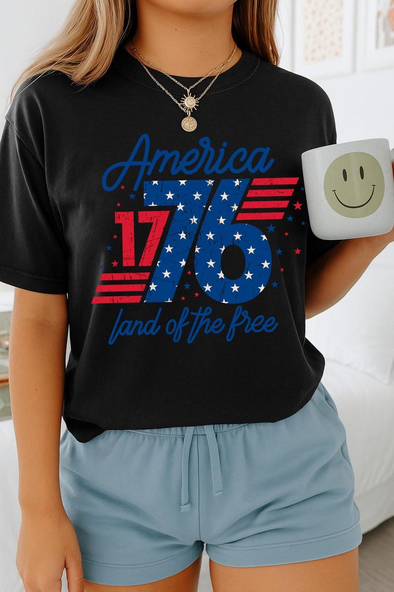 May include: Black t-shirt with the text "America 1776 land of the free" in red and blue. A white mug with a smiley face is held in the hand. The person is wearing blue shorts.
