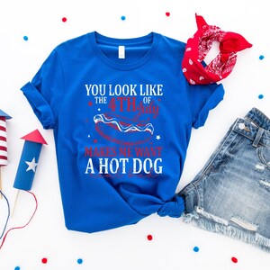May include: A royal blue t-shirt with the text "YOU LOOK LIKE THE 4TH OF JULY MAKES ME WANT A HOT DOG" in white and red. The image includes denim shorts, a red bandana, and decorative firework rockets.