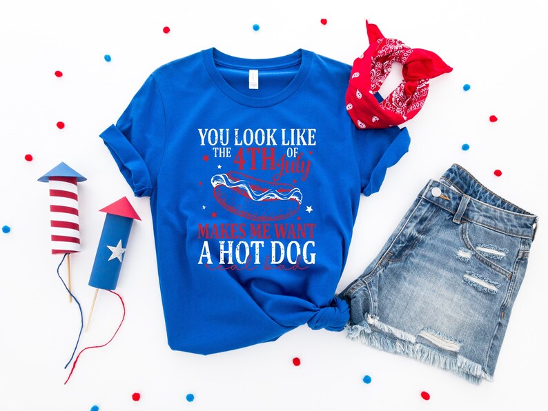 May include: A royal blue t-shirt with the text "YOU LOOK LIKE THE 4TH OF JULY MAKES ME WANT A HOT DOG" in white and red. The image includes denim shorts, a red bandana, and decorative firework rockets.