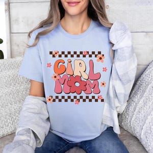 May include: Light blue t-shirt with a retro-style graphic that says "Girl Mom" in pink, orange, and yellow letters with a flower design. The graphic is surrounded by a black and white checkered border.