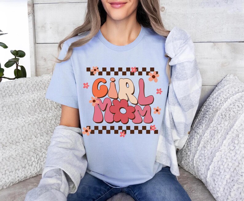 May include: Light blue t-shirt with a retro-style graphic that says "Girl Mom" in pink, orange, and yellow letters with a flower design. The graphic is surrounded by a black and white checkered border.