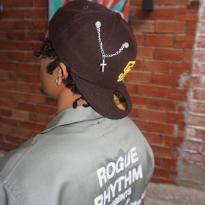 Könnte beinhalten: Eine Person trägt eine braune Baseballkappe mit einer silbernen Kette und einem Kreuzanhänger. Die Kappe hat ein gelbes Logo an der Seite. Die Person trägt ein grünes Hemd mit dem Aufdruck "Rogue Rhythm Presents" auf dem Rücken.