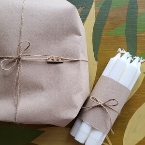 May include: A wrapped package and a bundle of white candles are presented on a decorative surface. The package is wrapped in brown paper and tied with twine. The candles are wrapped in brown paper and tied with twine.