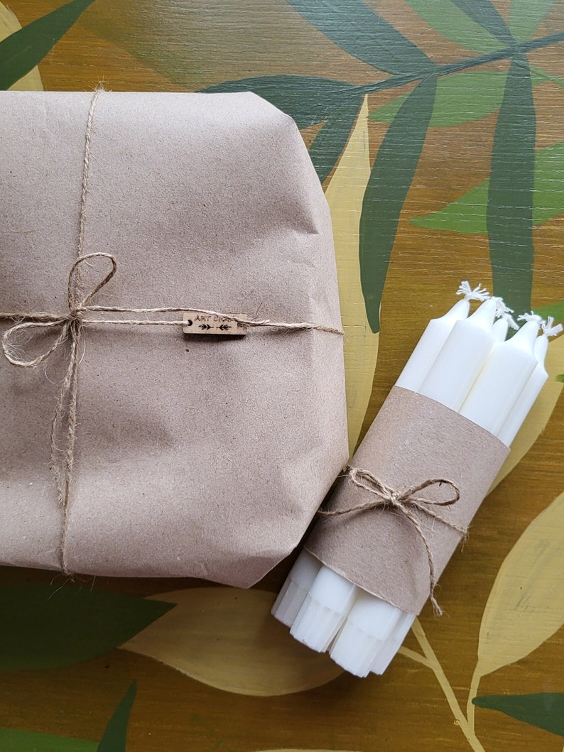 May include: A wrapped package and a bundle of white candles are presented on a decorative surface. The package is wrapped in brown paper and tied with twine. The candles are wrapped in brown paper and tied with twine.