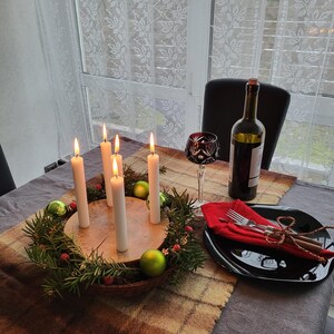 May include: A festive table setting with a lit Advent wreath centerpiece. The wreath is made of greenery, berries, and green ornaments. Tall white candles are lit. A wine bottle, a glass, and a black plate with silverware complete the scene.