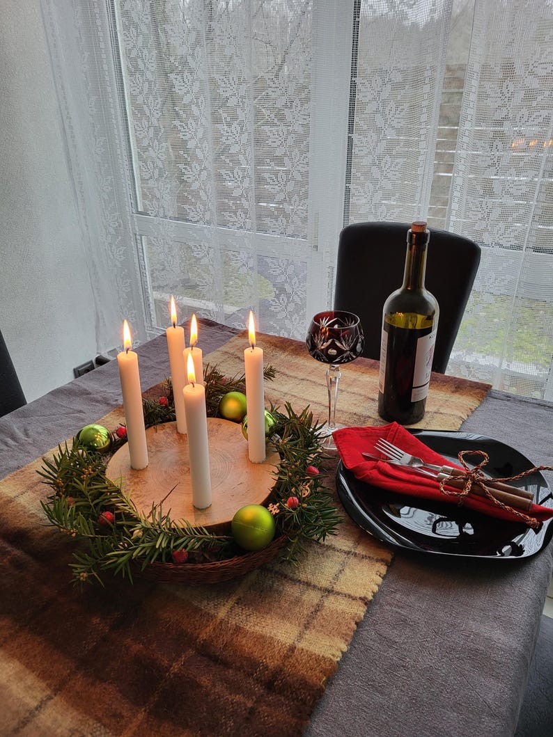 May include: A festive table setting with a lit Advent wreath centerpiece. The wreath is made of greenery, berries, and green ornaments. Tall white candles are lit. A wine bottle, a glass, and a black plate with silverware complete the scene.