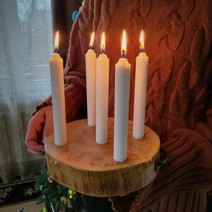May include: A wooden candle holder with six white candles, each with a lit flame. The holder is round, made from a tree cross-section, and has a natural wood grain. The candles are evenly spaced around the center.