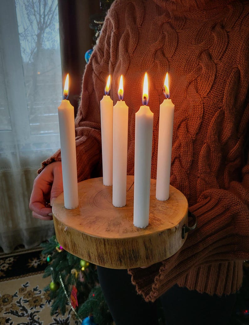 May include: A wooden candle holder with six white candles, each with a lit flame. The holder is round, made from a tree cross-section, and has a natural wood grain. The candles are evenly spaced around the center.