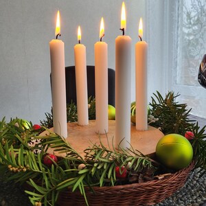May include: A festive arrangement featuring five lit white candles of varying heights. The candles are set on a wooden base, surrounded by green foliage, red berries, and green ornaments, all within a woven basket.