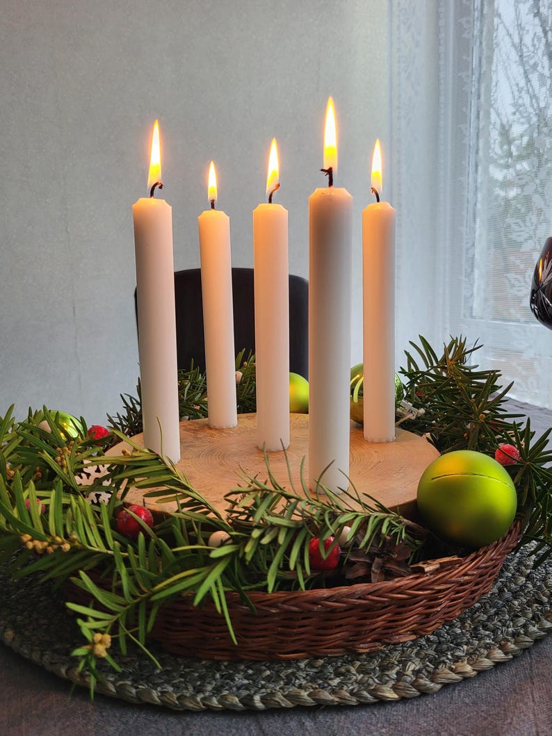 May include: A festive arrangement featuring five lit white candles of varying heights. The candles are set on a wooden base, surrounded by green foliage, red berries, and green ornaments, all within a woven basket.