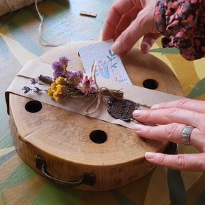 May include: A round, wooden gift box with a handle, wrapped in brown paper and twine. It is adorned with dried flowers and a wax seal. A small card with a floral design is placed on top.