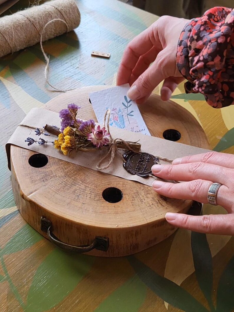 May include: A round, wooden gift box with a handle, wrapped in brown paper and twine. It is adorned with dried flowers and a wax seal. A small card with a floral design is placed on top.
