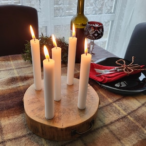 May include: A rustic centerpiece featuring six white candles with lit flames, arranged on a natural wood base. A wine bottle and a decorative glass are also on the table, set on a plaid tablecloth. The scene evokes a warm, inviting atmosphere.