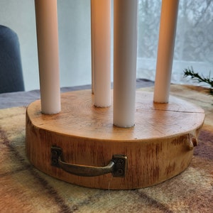 May include: A wooden candle holder with five white candles. The holder is made from a cross-section of a tree trunk, with a metal handle attached. The candles are lit, with flames visible.