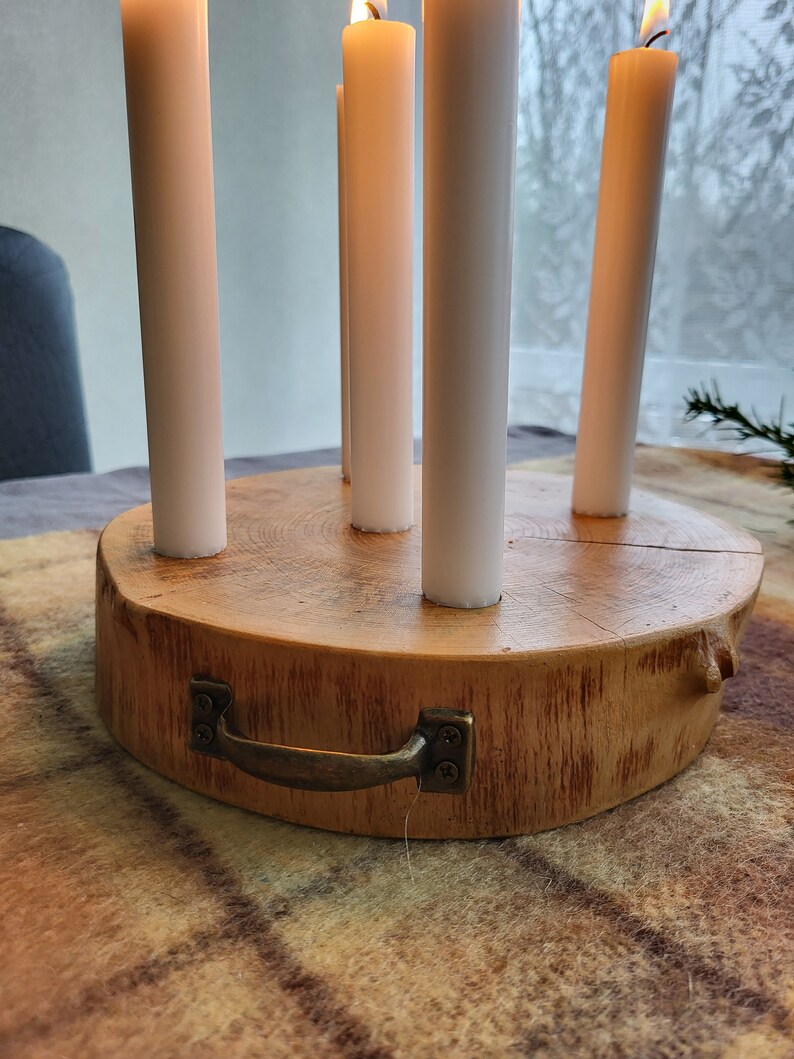 May include: A wooden candle holder with five white candles. The holder is made from a cross-section of a tree trunk, with a metal handle attached. The candles are lit, with flames visible.