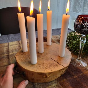 May include: A wooden candle holder with six white candles, each with a lit flame. The candles are arranged in a circle on a round, natural wood base. A decorative wine glass is visible in the background.