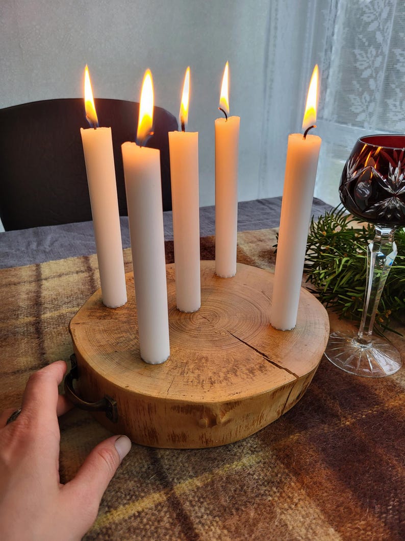 May include: A wooden candle holder with six white candles, each with a lit flame. The candles are arranged in a circle on a round, natural wood base. A decorative wine glass is visible in the background.