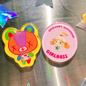 May include: Two resin-based pins. One is a yellow, orange, and purple bear-like character with an "X" on each cheek. The other is a pink circle with a cartoon character and the words "GASLIGHT GATEKEEP GIRLBOSS" in red.