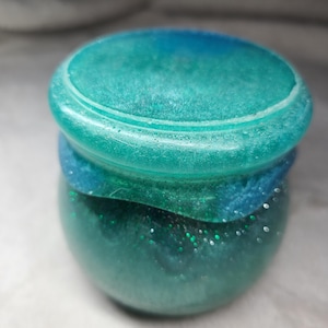 May include: A small, round jar with a lid, featuring a gradient of teal and blue hues. The jar's contents shimmer with green glitter. The lid has a raised edge, and the jar appears to be made of resin or a similar material.