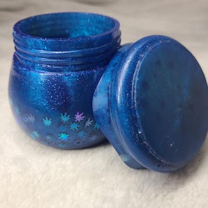 Könnte beinhalten: Ein blaues Glasgefäß mit Deckel, verziert mit schimmerndem Glitzer und holografischen Marihuana-Blatt-Akzenten. Das Gefäß ist rund und hat eine glatte, glänzende Oberfläche.