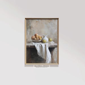 Puede incluir: Una pintura de bodegón que representa una jarra blanca, un cuenco de fruta y un paño blanco drapeado sobre una mesa de madera. El fondo es una pared beige texturizada. La pintura está enmarcada en un marco dorado.