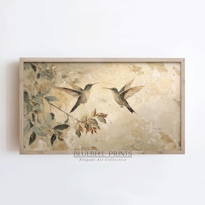 May include: A painting of two hummingbirds in flight against a beige background with green and brown foliage. The painting is framed in a light wood frame. The text "BLUEBELL PRINTS Elegant Art Collective" is at the bottom of the painting.