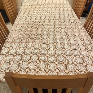 May include: A rectangular wooden dining table covered with a cream-colored, intricately patterned lace tablecloth. The table is surrounded by wooden chairs. The tablecloth features a repeating floral design.