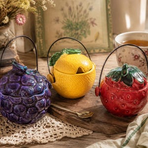 May include: Three ceramic lidded jars shaped like fruit, including a purple grape cluster, a yellow lemon, and a red strawberry. Each jar features a glossy finish, a removable lid with a green stem handle, and a thin black metal swing handle.