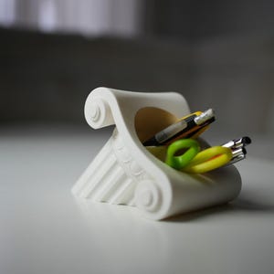 Pode incluir: Um organizador de secretária branco em forma de capitel de coluna clássica. Contém canetas e um elástico verde e amarelo. O design apresenta volutas e caneluras detalhadas. Um objeto decorativo e funcional.