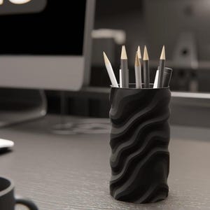 以下が含まれることがあります： グレーの机の上に、波打つ黒い鉛筆立てが置かれています。鉛筆立ての中には、鉛筆やペンがいくつか入っています。