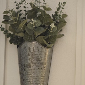 Może przedstawiać: Metalowa doniczka z vintage'owym wzorem, na której widnieje napis "Je t'aime" i inne francuskie frazy. Doniczka jest wypełniona bukietem zielonych, liściastych roślin z białymi kwiatami.