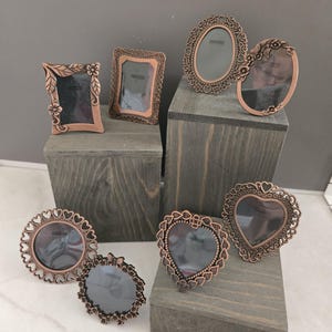 Puede incluir: Una colección de marcos de fotos decorativos de color cobre en varias formas, incluyendo ovalados, de corazón y rectangulares. Algunos marcos presentan adornos florales o en forma de corazón. Los marcos se exhiben sobre bloques de madera.