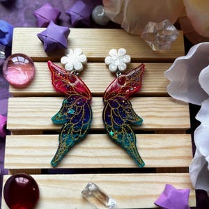 May include: A pair of iridescent butterfly wing earrings with gold glitter accents. The earrings are made of resin and have a white flower charm at the top.