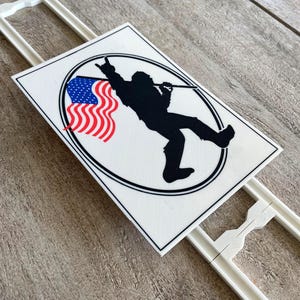 May include: A rectangular white sign featuring a black circular border. Inside the circle is a black silhouette of a person making a rock and roll hand gesture while holding a pole attached to a waving American flag with red, white, and blue colors.