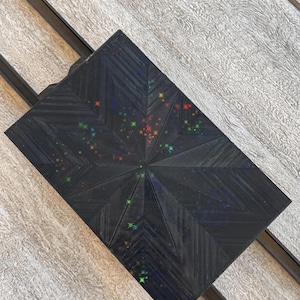 May include: A rectangular black box featuring a textured, geometric starburst pattern radiating from the center. The surface is adorned with small, scattered, multi-colored holographic star shapes in shades of red, green, blue, and yellow.