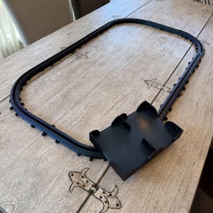 May include: A black plastic oval train track assembly featuring a rectangular platform section with raised dividers. The track is composed of multiple interlocking segments forming a continuous loop, designed for small-scale model train movement.