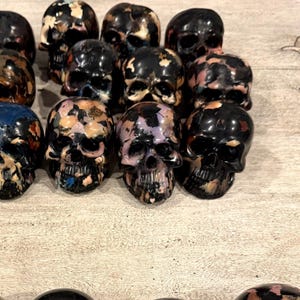 Peut inclure: Une collection de petites figurines de cr&acirc;nes en trois dimensions dispos&eacute;es en rang&eacute;es. Chaque cr&acirc;ne pr&eacute;sente une base noire brillante avec des &eacute;claboussures de peinture irr&eacute;guli&egrave;res et multicolores en bleu, rose, orange et blanc.