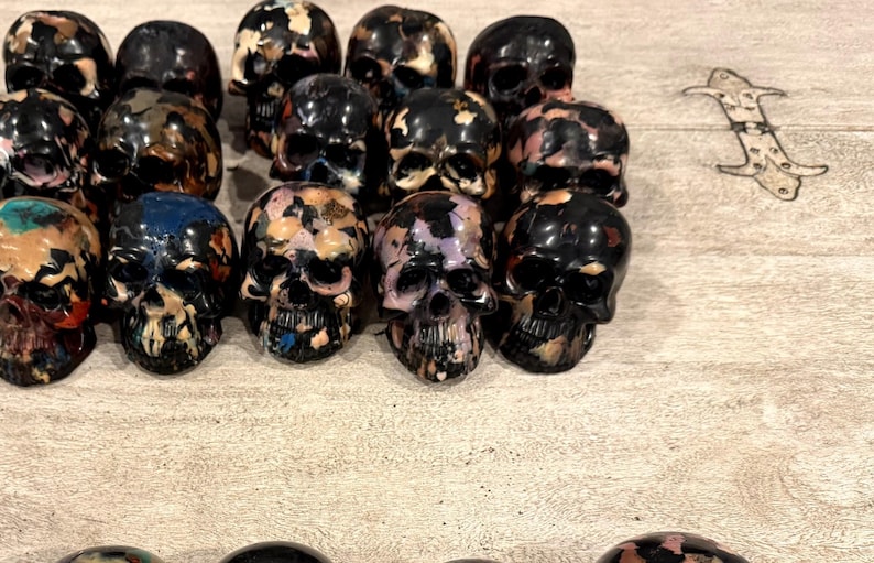 Peut inclure: Une collection de petites figurines de cr&acirc;nes en trois dimensions dispos&eacute;es en rang&eacute;es. Chaque cr&acirc;ne pr&eacute;sente une base noire brillante avec des &eacute;claboussures de peinture irr&eacute;guli&egrave;res et multicolores en bleu, rose, orange et blanc.