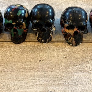 Peut inclure: Cinq objets en forme de cr&acirc;ne dispos&eacute;s en rang&eacute;e. Chaque cr&acirc;ne pr&eacute;sente une base noire brillante avec des &eacute;claboussures de peinture multicolores uniques dans des tons de rose, bleu, vert et orange. Les cr&acirc;nes ont des orbites et des cavit&eacute;s nasales creuses.