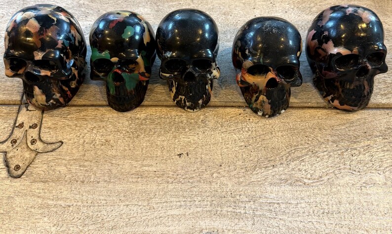 Peut inclure: Cinq objets en forme de cr&acirc;ne dispos&eacute;s en rang&eacute;e. Chaque cr&acirc;ne pr&eacute;sente une base noire brillante avec des &eacute;claboussures de peinture multicolores uniques dans des tons de rose, bleu, vert et orange. Les cr&acirc;nes ont des orbites et des cavit&eacute;s nasales creuses.