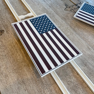 May include: Two white plastic frames with a red, white, and blue American flag design on a wooden surface. The flag is on a track and is sliding through the frame.