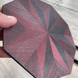 May include: An octagonal coaster with a geometric starburst design. The coaster features a pattern of burgundy and gray triangular shapes radiating from the center. The coaster is held by a hand.