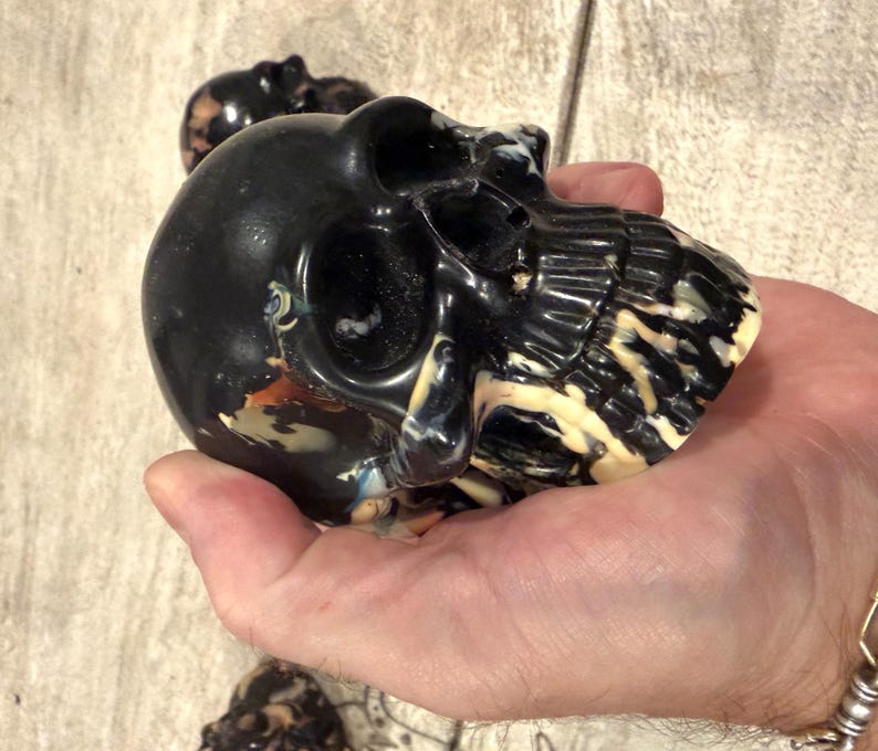 Peut inclure: Une sculpture de cr&acirc;ne noire pr&eacute;sentant des tourbillons marbr&eacute;s de couleurs cr&egrave;me, beige et bleu sur toute la surface. Le cr&acirc;ne poss&egrave;de des orbites, une cavit&eacute; nasale et des dents d&eacute;taill&eacute;es. Un second cr&acirc;ne plus petit est visible en arri&egrave;re-plan.