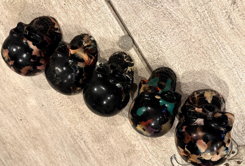 Peut inclure: Cinq sculptures de cr&acirc;nes polies dispos&eacute;es en ligne diagonale, chacune pr&eacute;sentant une base noire brillante avec des inclusions multicolores beige, blanc et sarcelle. Une pi&egrave;ce de monnaie est plac&eacute;e entre le deuxi&egrave;me et le troisi&egrave;me cr&acirc;ne.