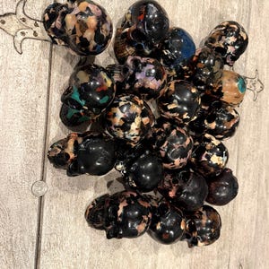 Peut inclure: Une collection de petites figurines en forme de cr&acirc;ne fabriqu&eacute;es &agrave; partir d'une r&eacute;sine sombre et multicolore. Chaque cr&acirc;ne pr&eacute;sente un motif mouchet&eacute; unique de noir, de beige, de blanc et de taches bleues ou vertes, dispos&eacute;s en groupe dense.