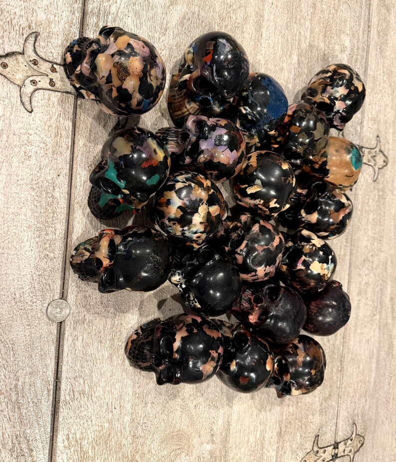 Peut inclure: Une collection de petites figurines en forme de cr&acirc;ne fabriqu&eacute;es &agrave; partir d'une r&eacute;sine sombre et multicolore. Chaque cr&acirc;ne pr&eacute;sente un motif mouchet&eacute; unique de noir, de beige, de blanc et de taches bleues ou vertes, dispos&eacute;s en groupe dense.