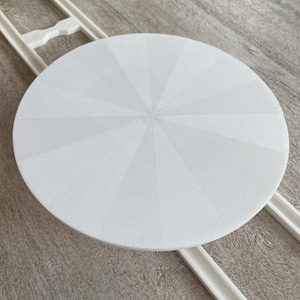 May include: A white, round cake stand with a textured surface and a sunburst design. The stand is supported by a white, rectangular frame. The stand is designed for displaying cakes and other baked goods.