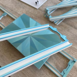 May include: A collection of light blue and white decorative frames and components. The frames have a geometric design with a white border and a light blue rope-like detail. Additional pieces include a white rectangular base and striped elements.